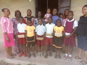 Comrd. (Engr.) Promise Chidiebere Ezekiel with his daughter, Miss Prestige Promise (M); Mrs. Precious Promise (2R); Mrs. Oriaku Uzoma Rose, the Admin Head Mistress (1R); Mrs. Omire Helene Onuabuchi, the assistant HM (1R), flanked by other teachers and pupils (beneficiaries).